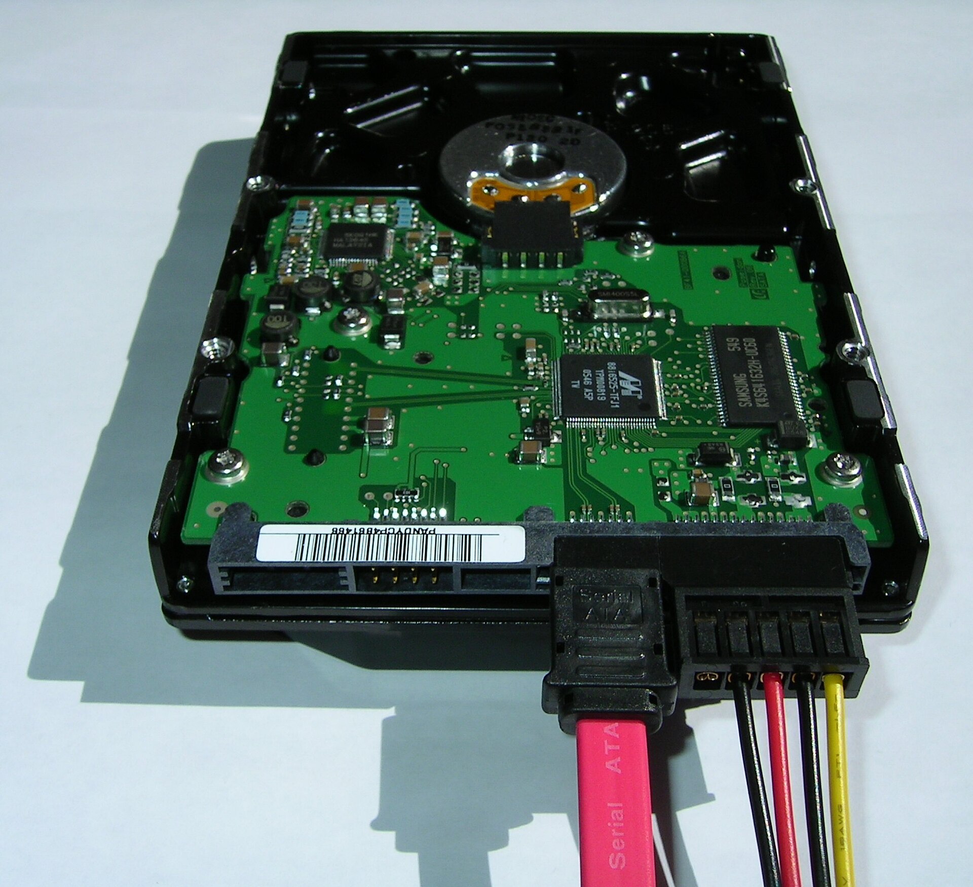 The underside of a 3.5-inch hard drive showing the green circuit board with a large controller chip, a memory chip and the SATA connectors with a red data cable and a power plug