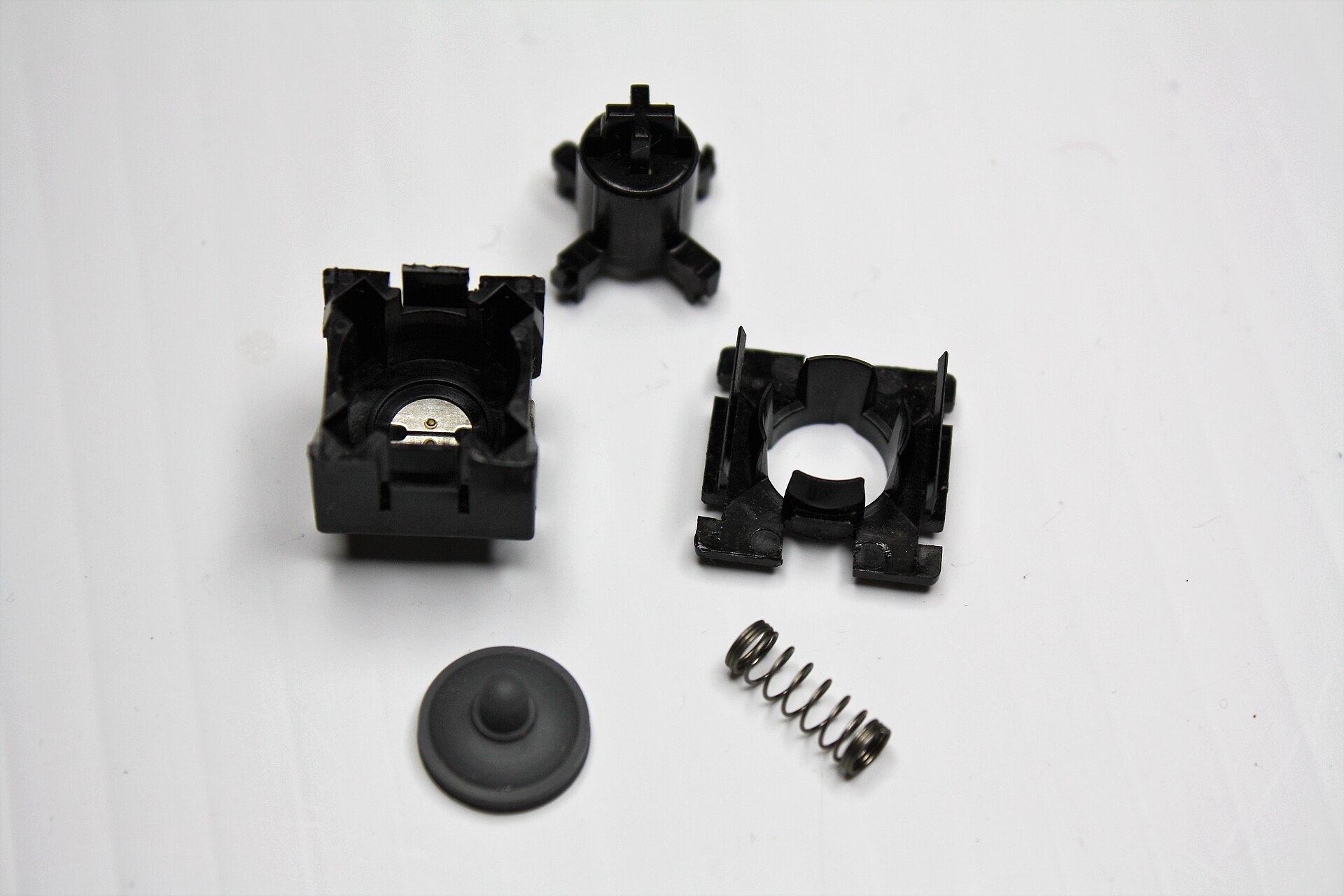 A single keyboard key switch taken apart on a white surface, showing a black plastic housing with a metal contact, a plunger with a cross-shaped stem, a square frame, a spring and a grey rubber dome-like part