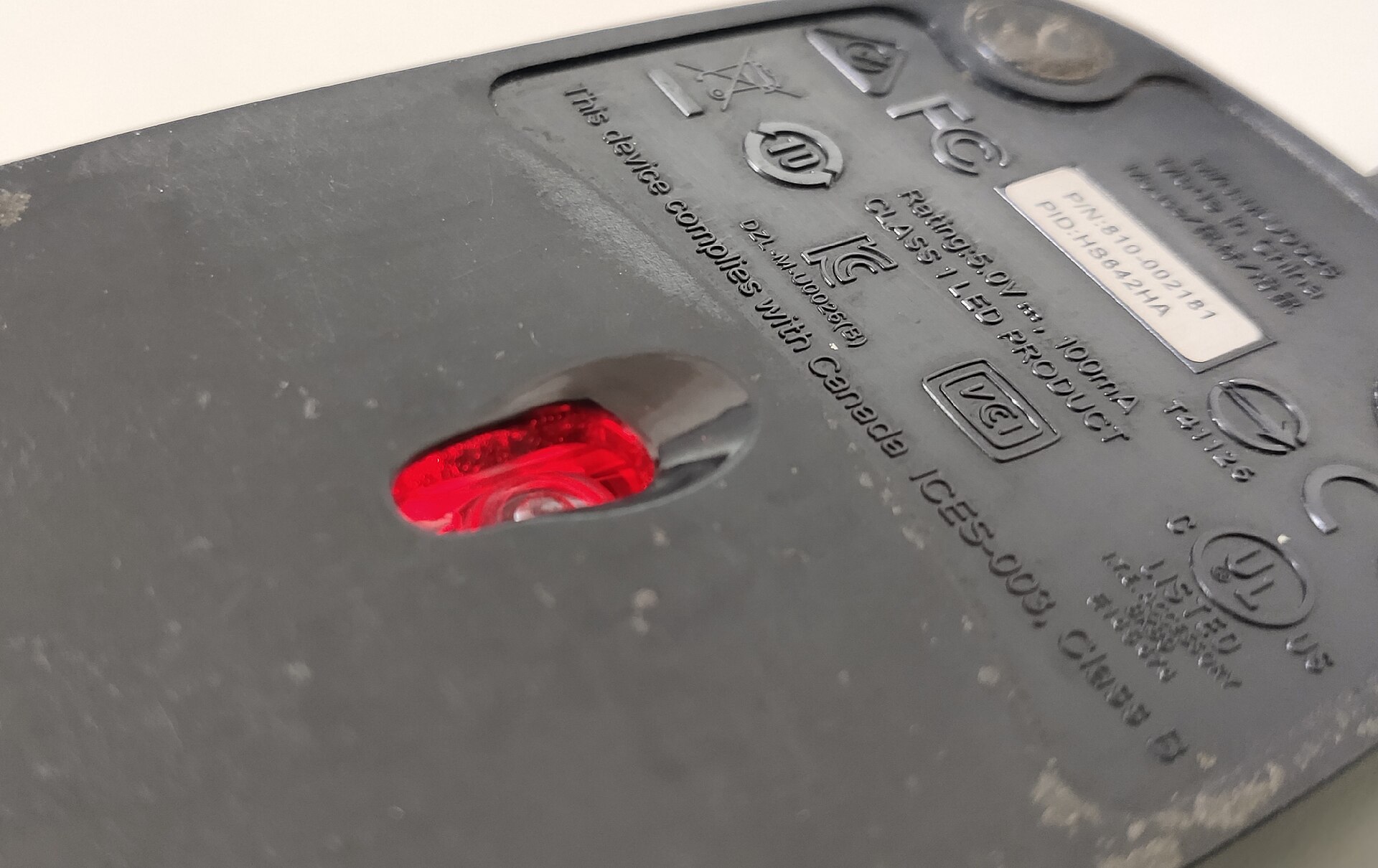 Underside of a black computer mouse showing a glowing red window where the optical sensor looks at the surface, plus regulatory markings